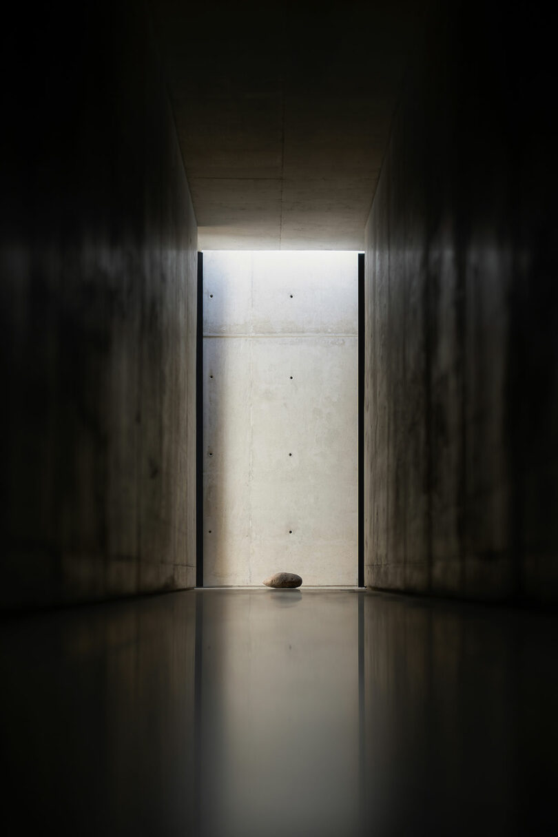 A single stone is placed on the floor at the end of a narrow, dimly lit concrete corridor with light entering from an opening above.
