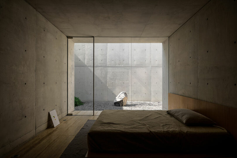 A minimalist bedroom with concrete walls, a low bed, and a glass door opening to a small, enclosed courtyard with a large rock and sparse gravel.