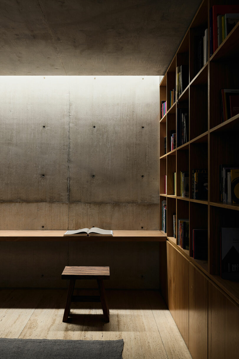 A minimalist reading nook features a wooden bench with an open book, a small wooden stool, built-in bookshelves, and a concrete wall with soft overhead lighting.