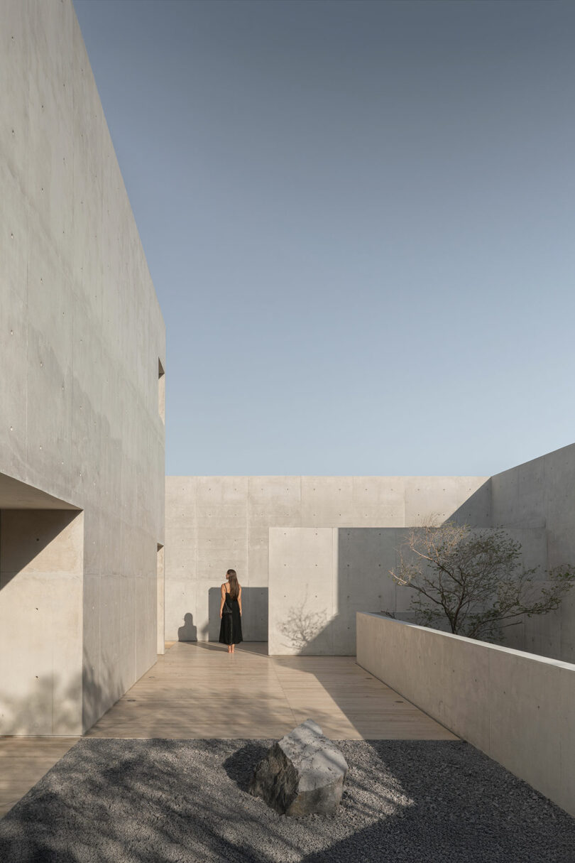 A woman in a black dress stands in a minimalist concrete courtyard with gravel, a large rock, and a small tree under a clear sky.