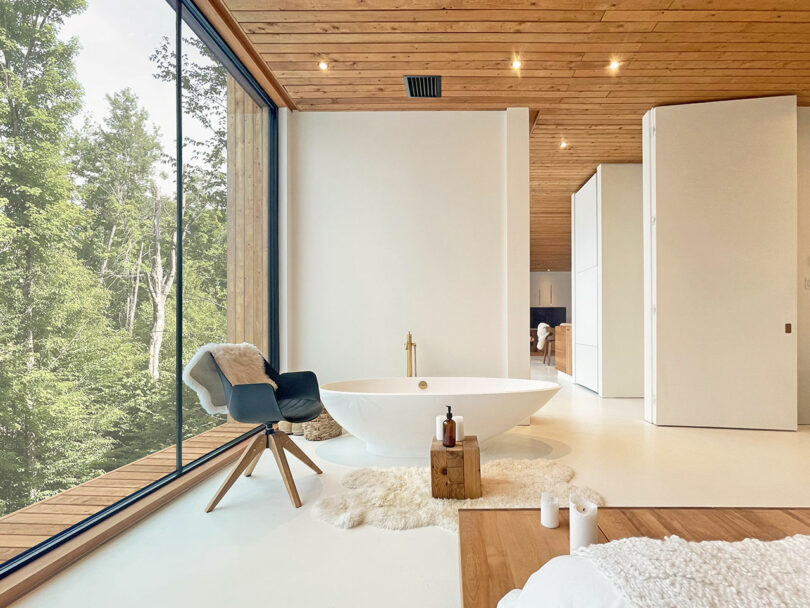 Modern bathroom with a freestanding tub, wooden stool, black chair, and floor-to-ceiling window overlooking trees; minimalist decor with white walls and ceiling.