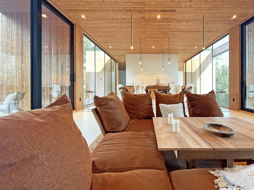 Modern living and dining area with large brown sectional sofa, wooden table, and minimalist decor, featuring floor-to-ceiling windows and wood-paneled ceiling and walls.