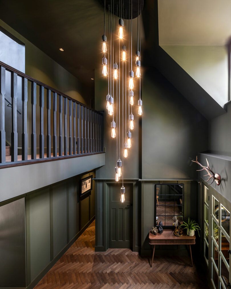 A modern entryway with dark green paneled walls, a cluster of hanging pendant lights, herringbone wood floor, a staircase, and a console table with plants and a mirror.
