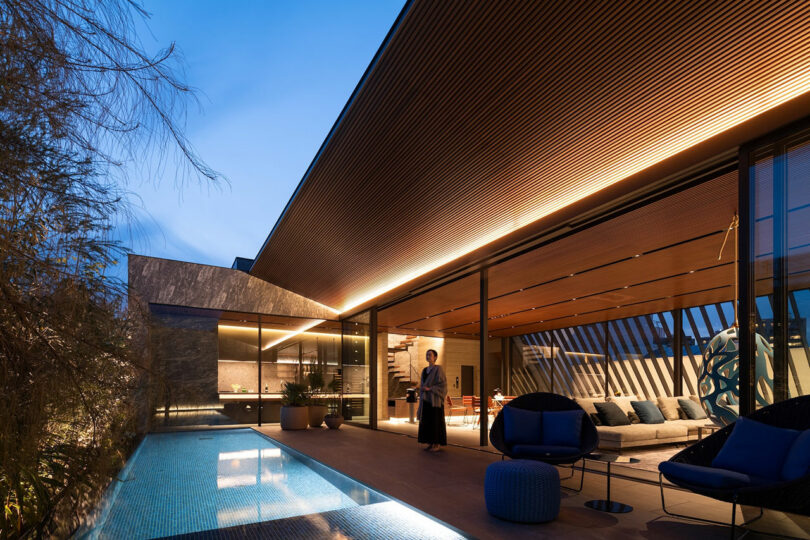 Modern house with large glass walls, a patio seating area, and a narrow illuminated pool at dusk. A person stands near the sliding doors, with warm lights inside and outside.