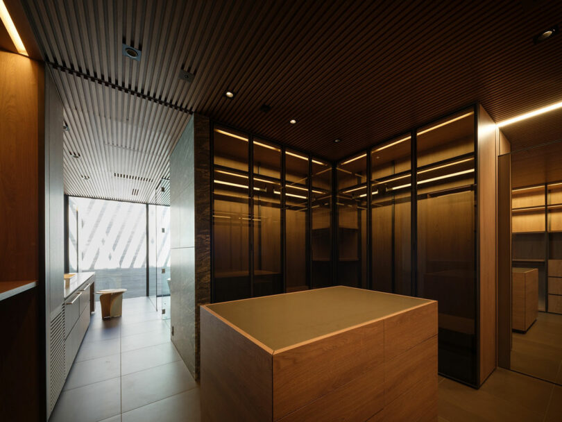 Modern office interior with wooden finishes, glass partitions, and minimalist furnishings, featuring a central counter and natural light from large windows.