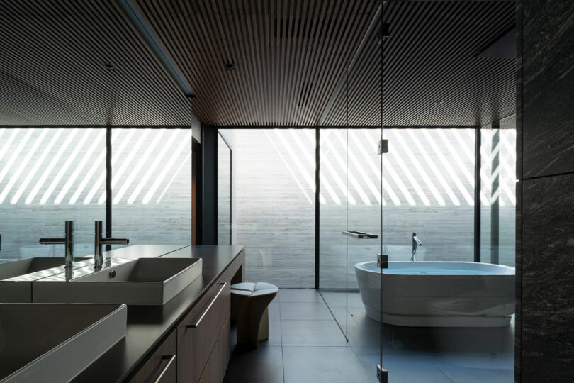 Modern bathroom with double sinks, freestanding bathtub, floor-to-ceiling windows with diagonal light patterns, and wood slat ceiling.