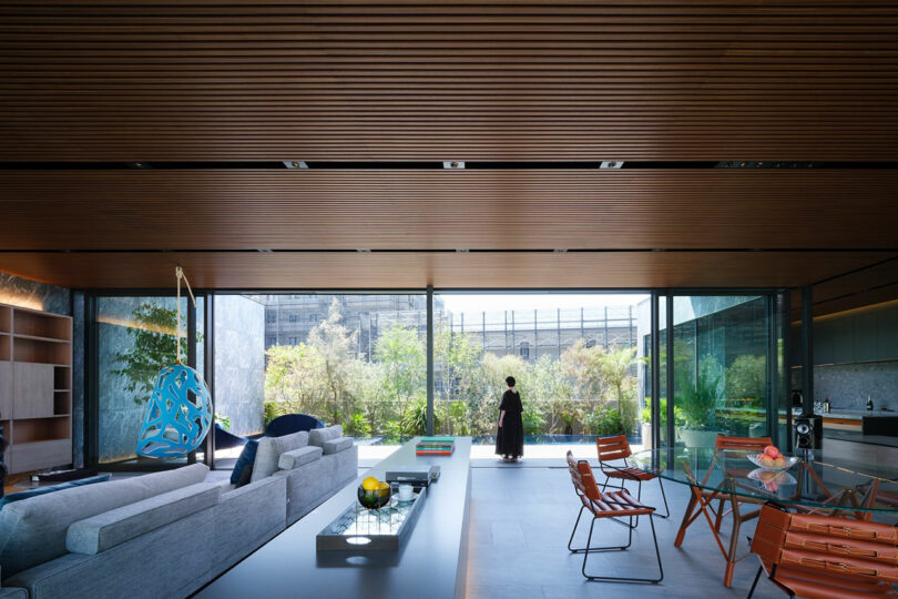 Spacious modern living room with floor-to-ceiling windows, wooden ceiling, contemporary furniture, and a person standing outside on the patio.