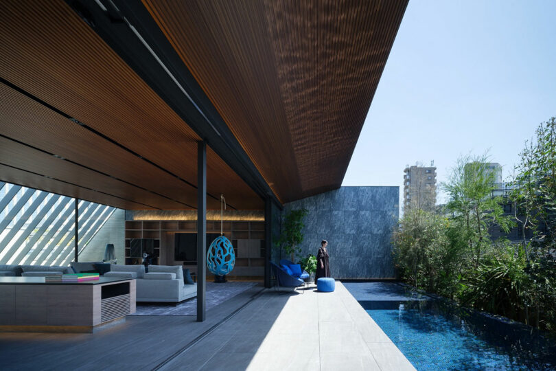 A modern open-plan living area with large overhanging roof, outdoor pool, and a person standing near greenery on a sunny day.