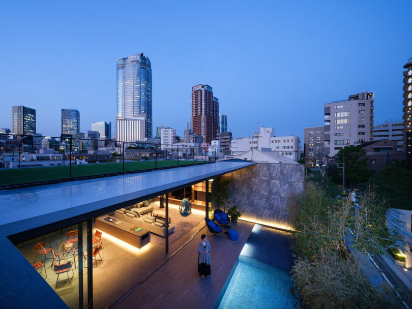 A modern rooftop terrace with seating, a pool, and city skyscrapers in the background at dusk; a person stands near the pool.