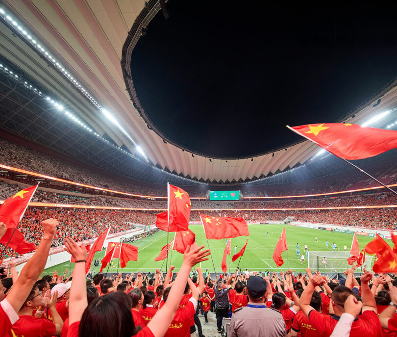 A large crowd in a stadium waves Chinese flags while watching a soccer match, with teams playing on the field under bright stadium lights.