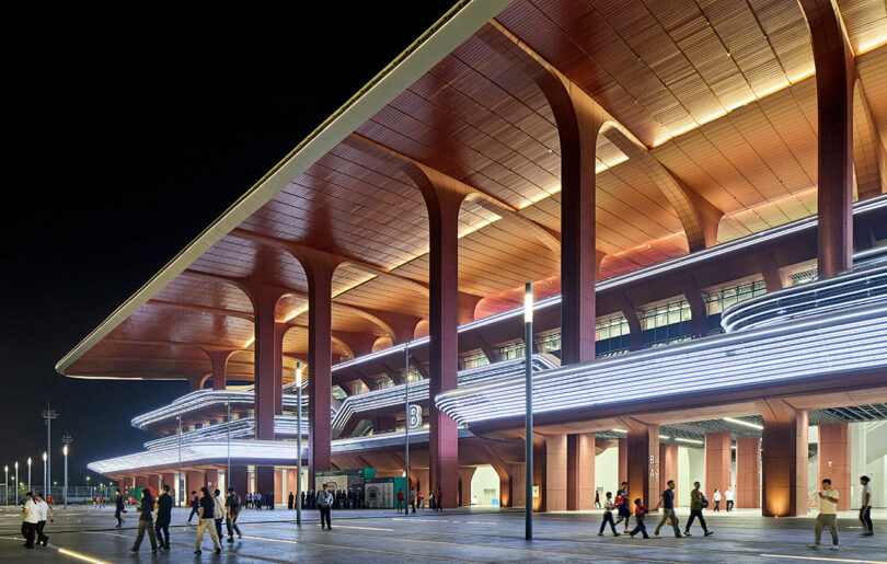 People walk outside a large, modern building with tall columns and illuminated horizontal lines at night.