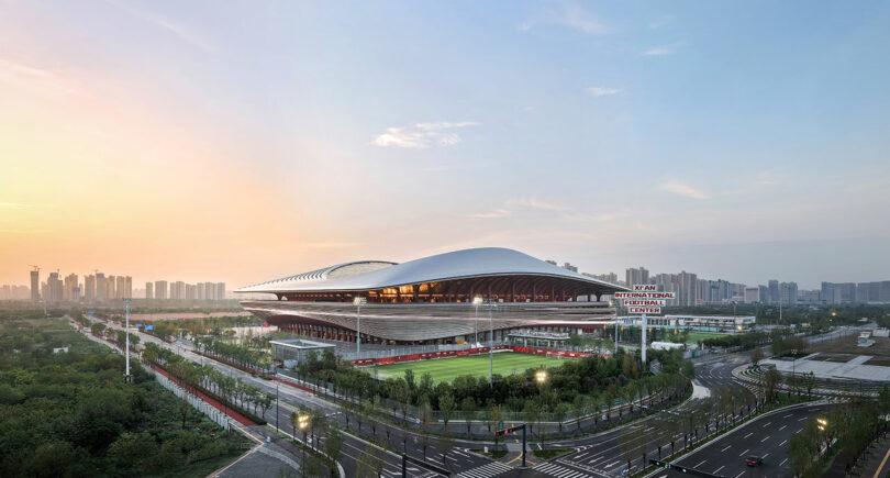 Large modern sports stadium with a unique curved roof, surrounded by green spaces and roads, set against a city skyline at sunset. A sign reads “Xiamen International Football Centre.”.