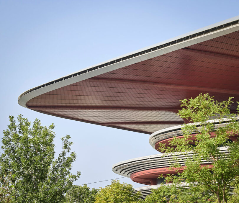 Large, futuristic building with layered, disc-shaped roofs above green trees, set against a clear blue sky.