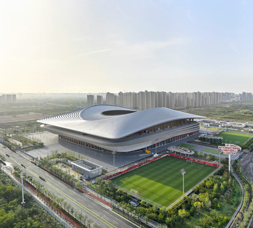 Aerial view of a large, modern stadium with a unique wavy roof design, adjacent to a soccer field, surrounded by greenery and city buildings in the background.