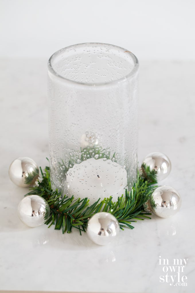 Decorative garland tie with small ornaments attached wrapped around a glass candle holder.