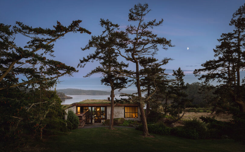 A small house with warm interior lighting sits among tall trees at dusk, with a view of water and distant hills under a partly cloudy sky and a crescent moon.
