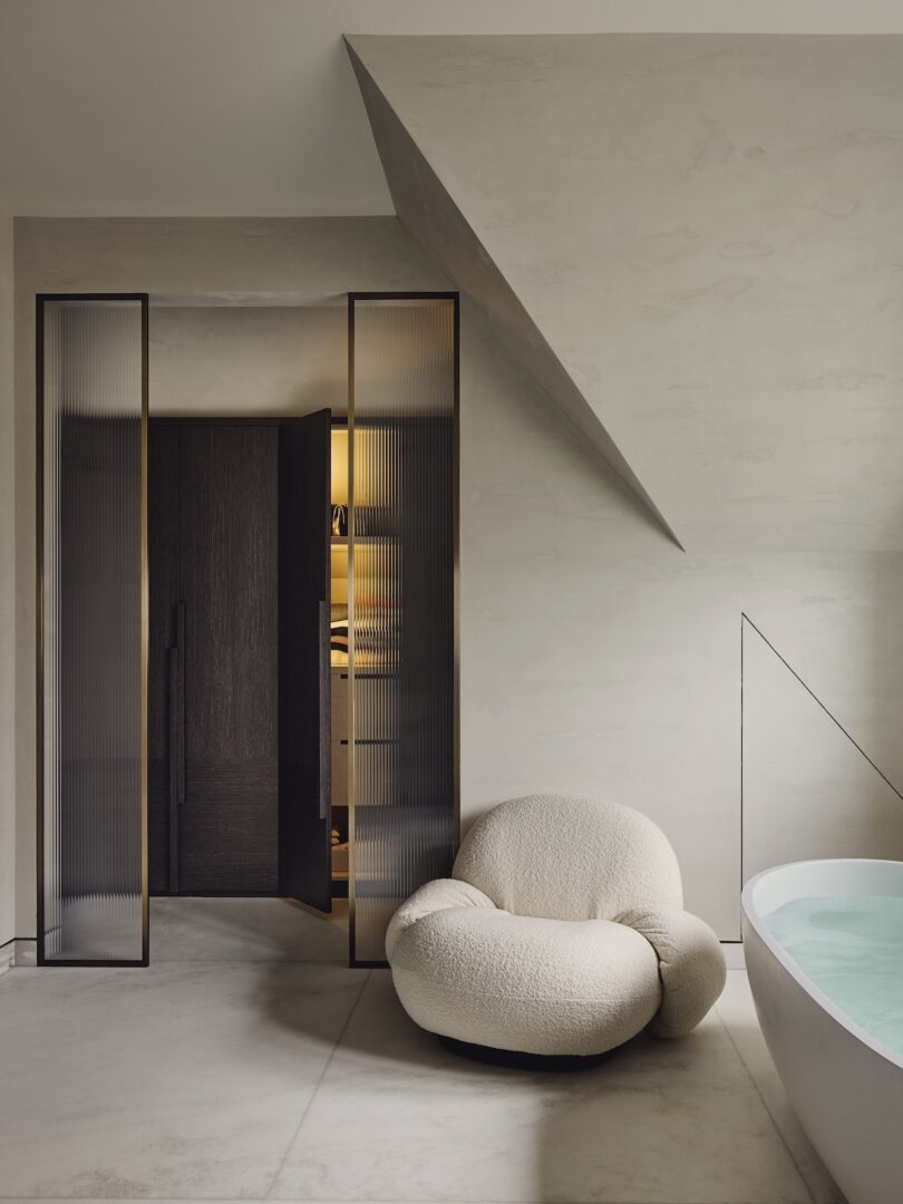 Minimalist bathroom with a textured cream lounge chair, glass double doors, built-in storage, and a white bathtub partially filled with water under sloped ceiling.