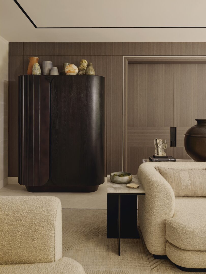 A modern living room with a curved beige sofa, a dark wood cabinet decorated with vases, neutral-toned walls, and minimalist decor.
