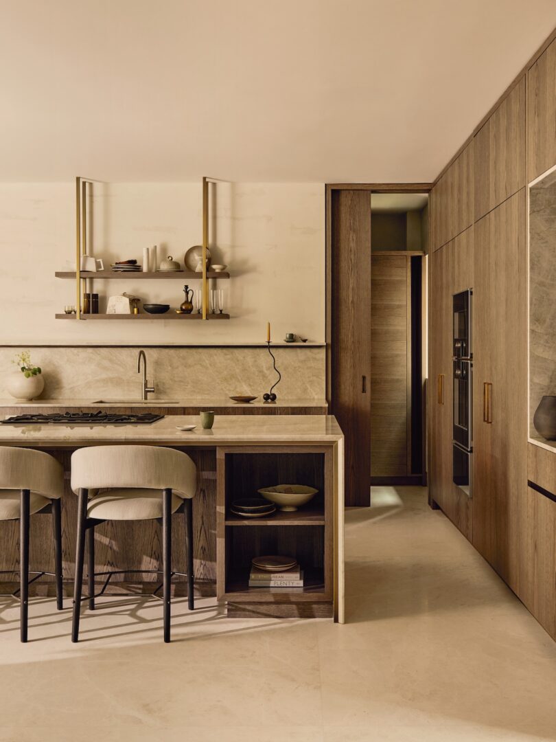 Modern kitchen with wood cabinetry, marble countertops, open shelves with pottery, built-in appliances, and two upholstered bar stools at the island.