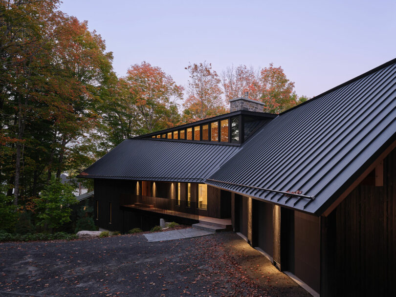 Modern house with dark metal roof and wood siding, surrounded by autumn trees at dusk, with warm interior lights visible through large windows.