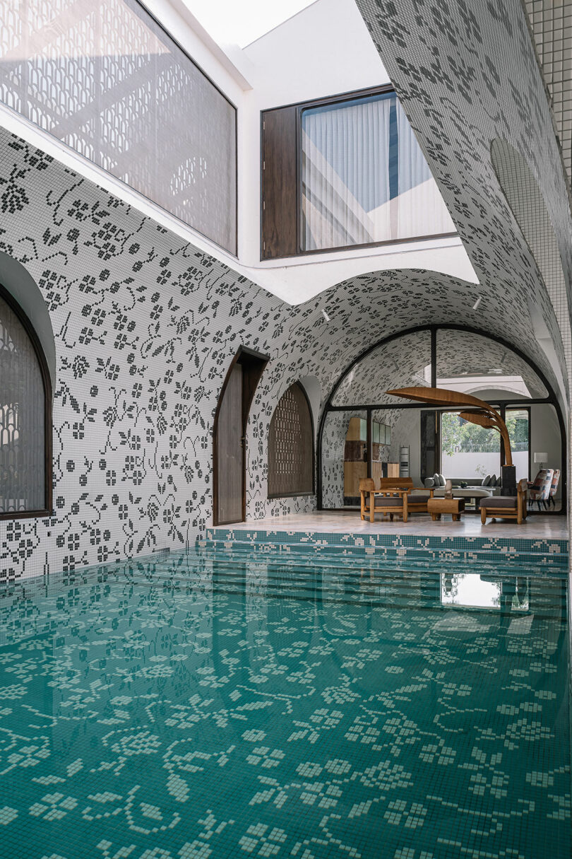 Indoor swimming pool with mosaic tile walls and ceiling in a floral pattern, arched doorways, and a lounge area with wooden furniture in the background.