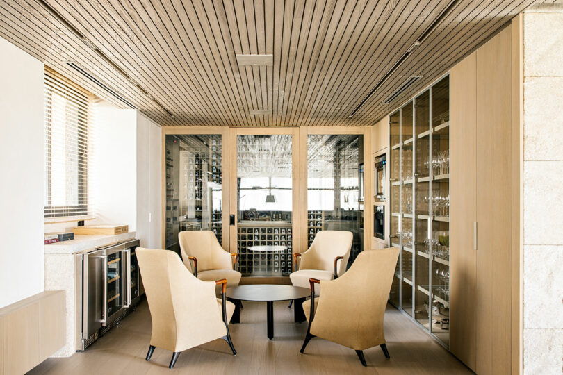 A modern lounge area with four beige armchairs around a round table, glass doors, a wine rack, and wooden ceiling panels.