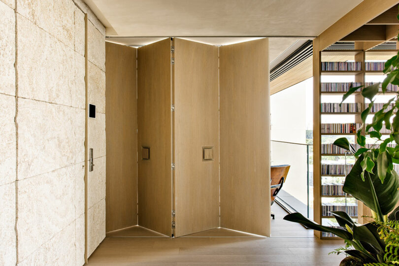 A folding wooden door partially open in a modern interior with stone and wood walls, shelves with books, and indoor plants.