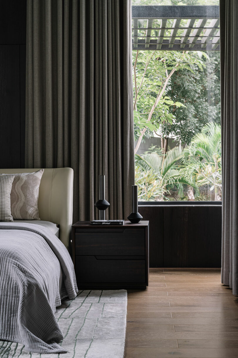 A modern bedroom with a bed, bedside table, and decor, featuring large windows with a view of lush greenery outside.