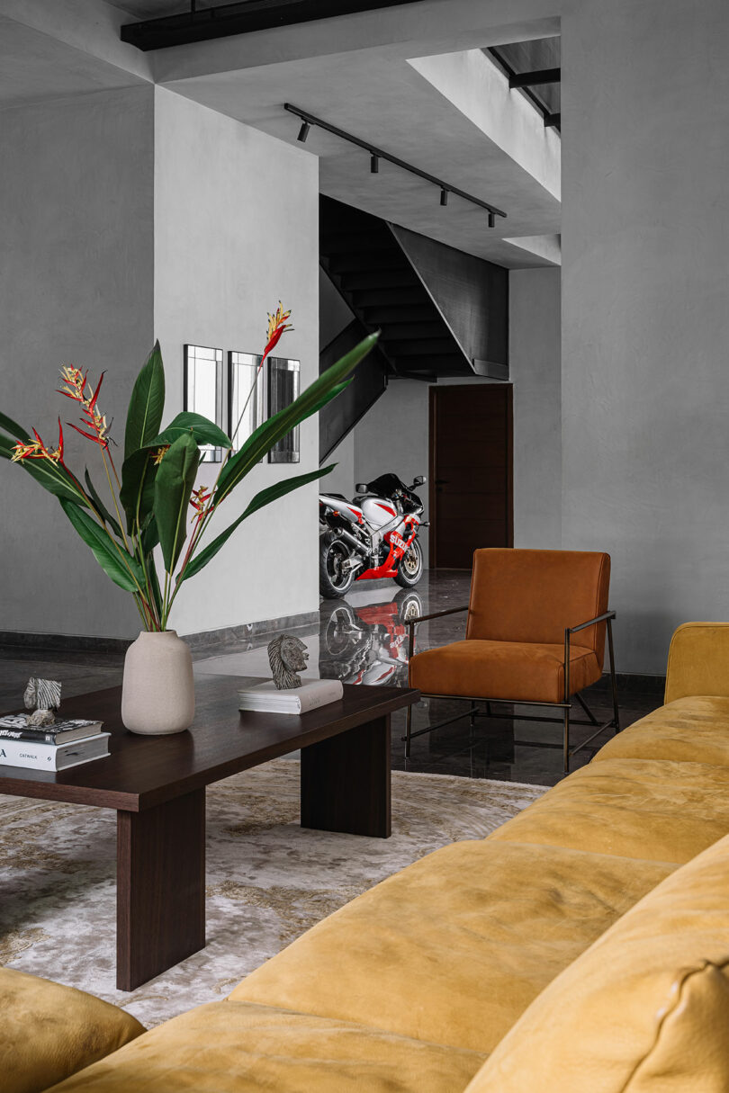 Modern living room with a yellow sofa, brown chair, and dark wood coffee table with a vase of flowers; a red and white motorcycle is visible in the hallway near a staircase.