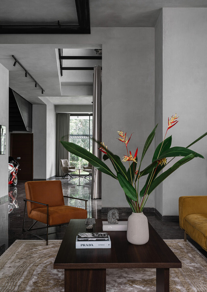 Modern living room with minimalist decor, featuring a brown armchair, yellow sofa, dark wood coffee table with a plant, and large windows allowing natural light. A motorcycle is visible in the background.